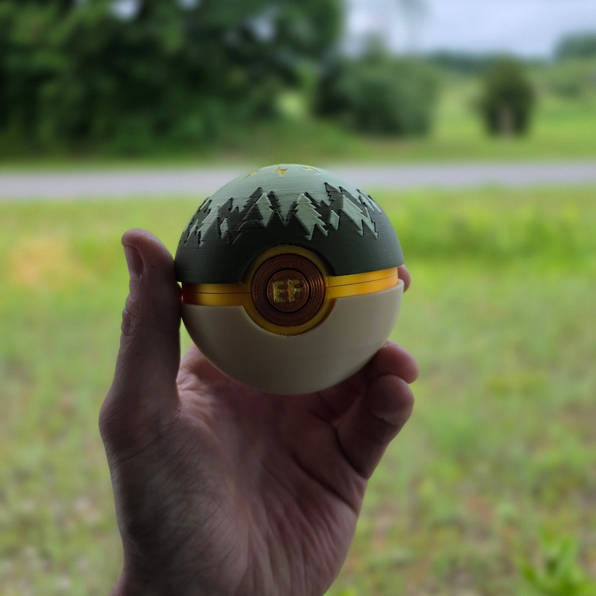 A hand holding a custom Pokeball-style container outdoors, featuring a forest tree silhouette pattern on the top half and a gold emblem on the bottom half, with a blurred green field and trees in the background.