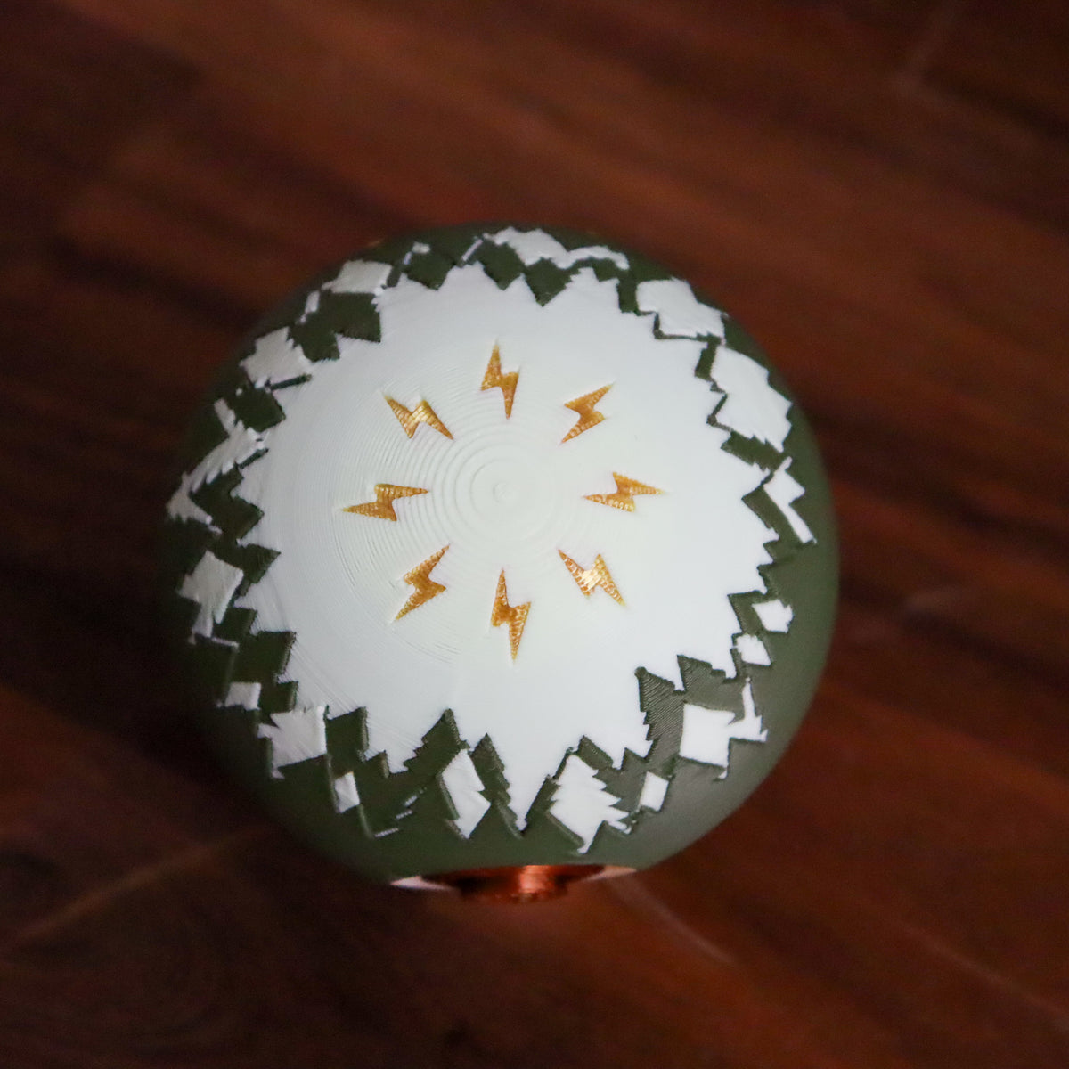 A spherical decorative object with a white top half featuring a gold lightning-bolt pattern, a dark olive-green lower half, and a jagged white border, resting on a dark wooden surface.