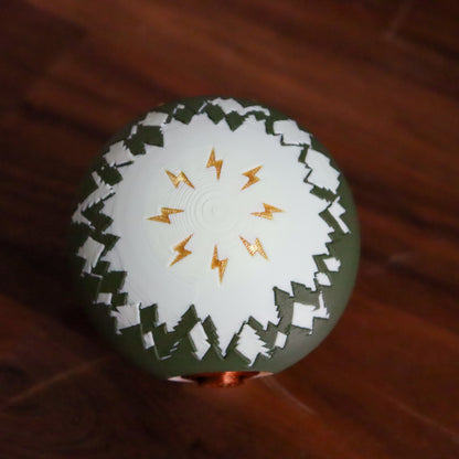 A spherical decorative object with a white top half featuring a gold lightning-bolt pattern, a dark olive-green lower half, and a jagged white border, resting on a dark wooden surface.