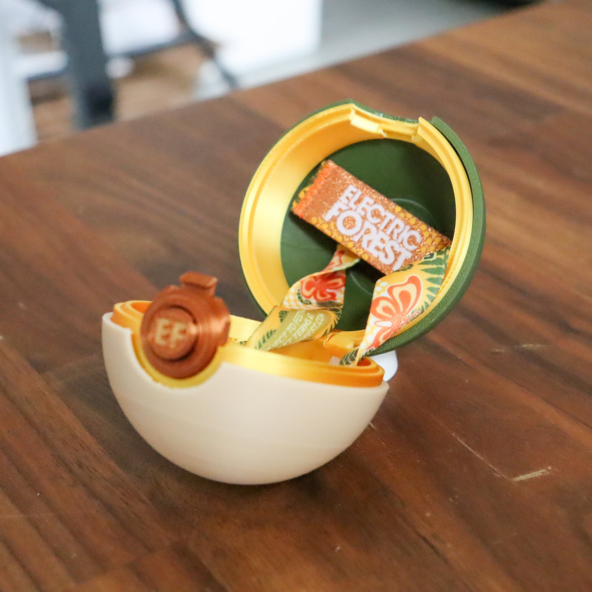 A small decorative egg-shaped container opened on a wooden table, with a white lower half, gold trim, a green inner lid, and a ribbon inside that reads 'Electric Forest.'.