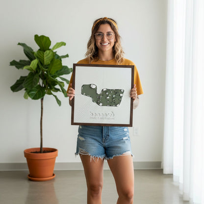 Woman holding a framed festival map of bonnaroo in  a room with a plant and white wall.