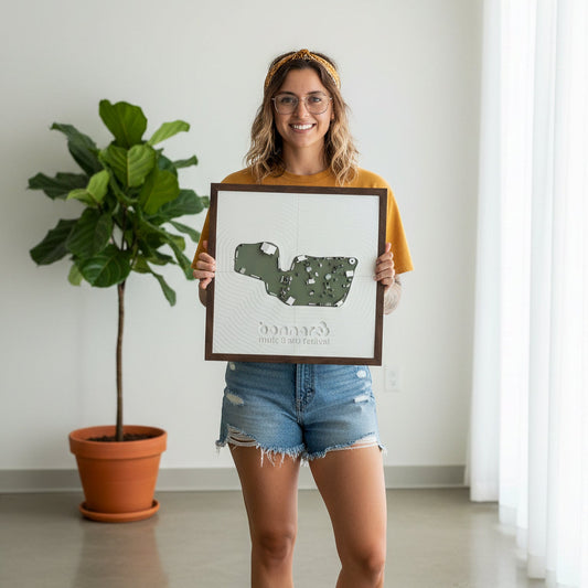 Woman holding a framed festival map of bonnaroo in  a room with a plant and white wall.