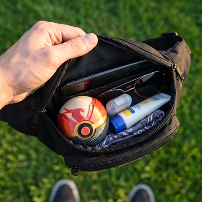 A black waist bag or fanny pack is shown open in an outdoor setting, held by a person's hand. Inside the bag are various items including a red and white spherical accessory, sunglasses, a tube of sunscreen, and a phone.