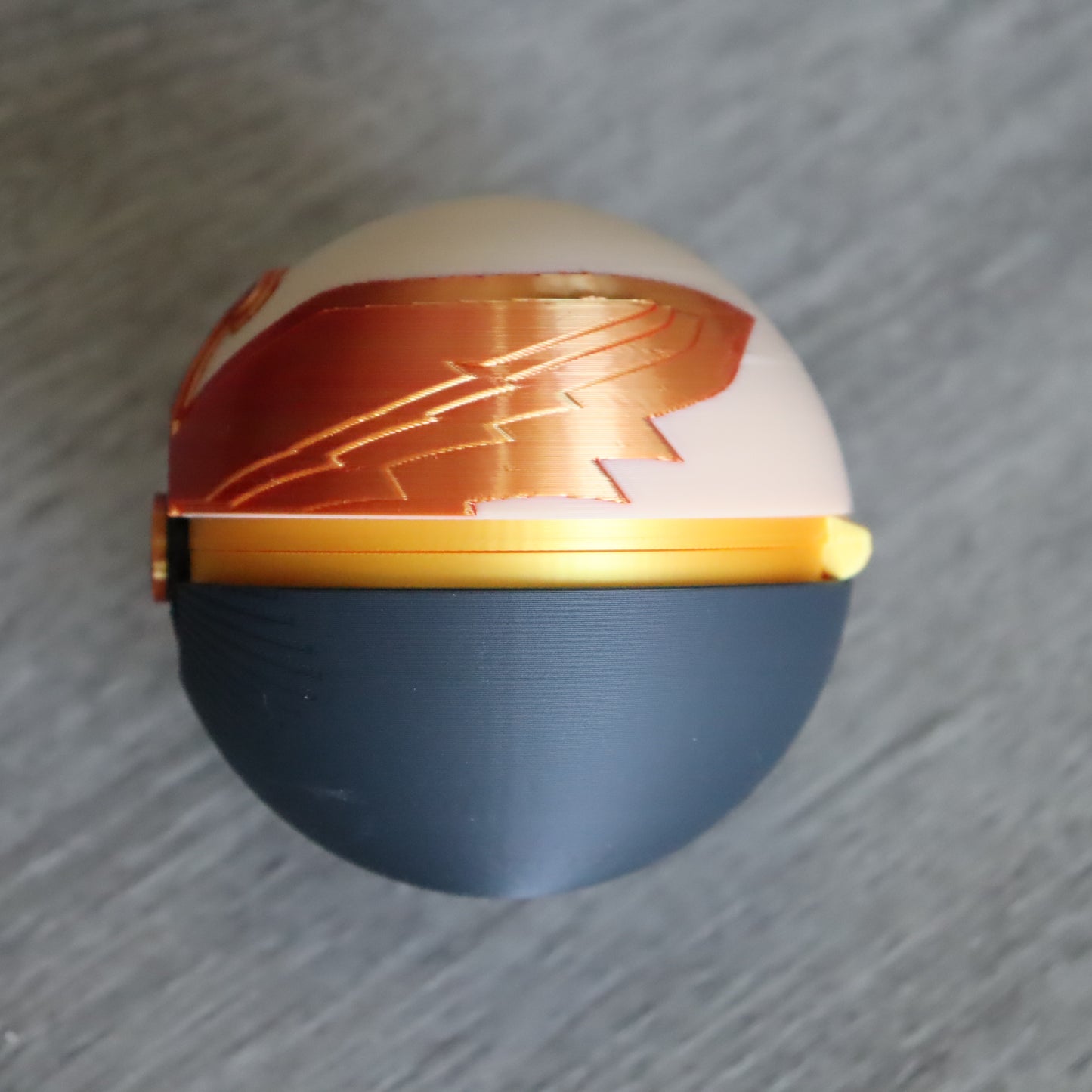 A spherical object with a split design, featuring a matte dark blue lower half and a glossy light beige upper half with a metallic copper-orange graphic pattern. A gold band encircles the middle, and a small gold clasp is visible on the left, set against a gray textured background.