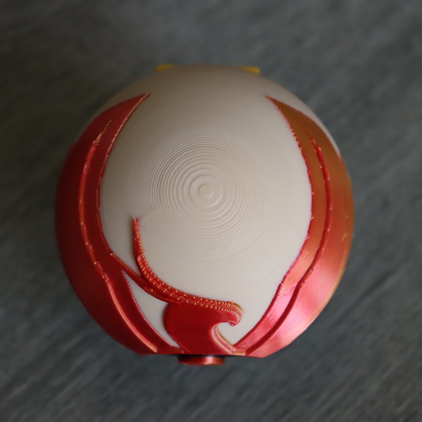 A spherical decorative object with a cream-colored top featuring concentric circular grooves and red sculpted side details, photographed on a gray textured surface.